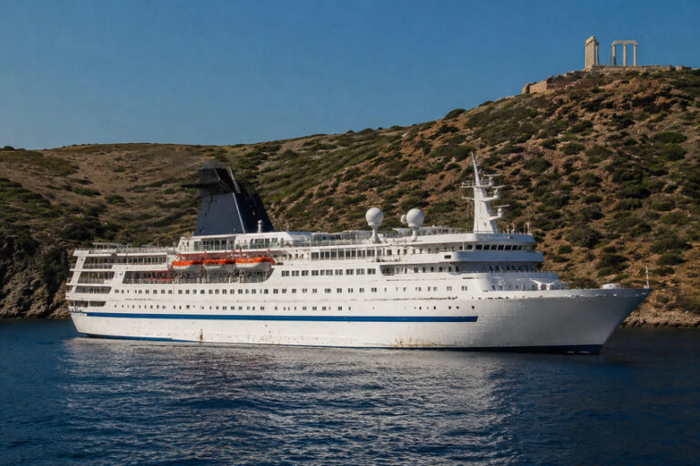 White cruise ship anchored along a rocky coastline, with an ancient ruin on the hilltop in the background.