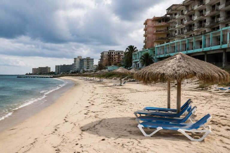 Row of blue lounge chairs under a thatched umbrella on a sandy, near-empty beach with unfinished hotel buildings along the shore under a cloudy sky, Caribbean feel.