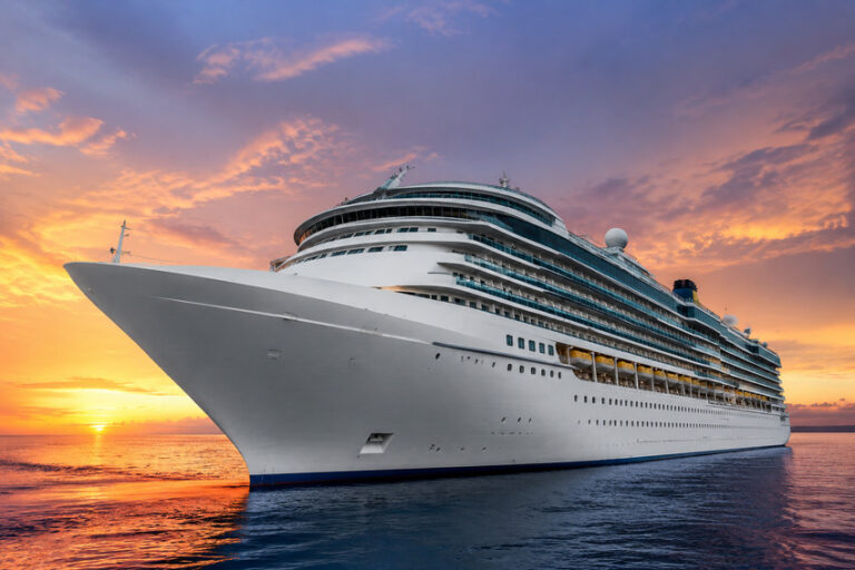 Large white cruise ship on calm ocean at sunset with an orange-pink sky behind it