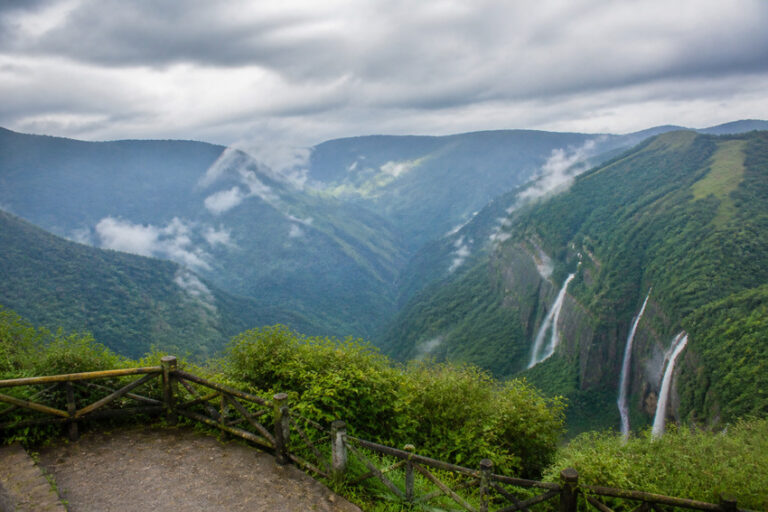 Scenic viewpoint over a green valley with multiple waterfalls on the right and misty clouds above, wooden railing in the foreground.