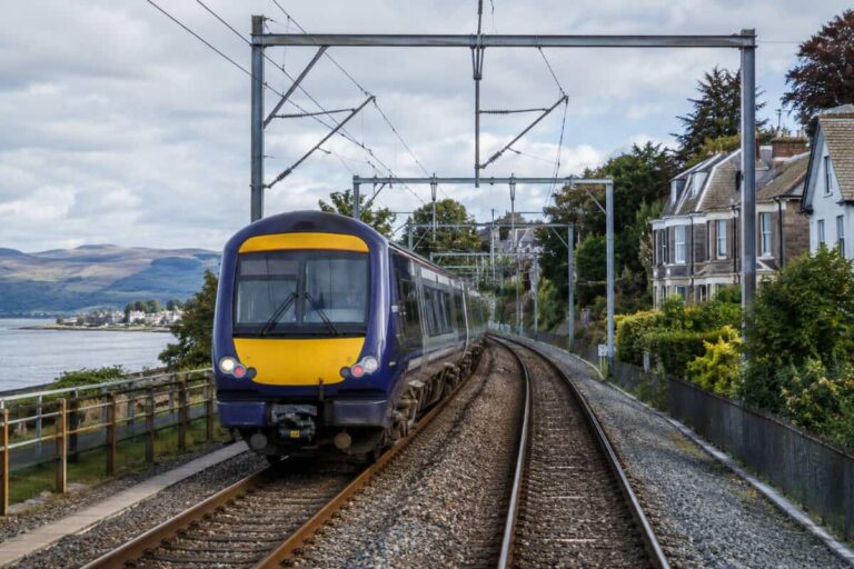 Blue and yellow passenger train on a curved coastal track with houses and hills in the background.