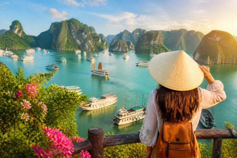 Woman wearing a conical hat and backpack overlooks a turquoise bay with boats and distant limestone karsts at sunset.