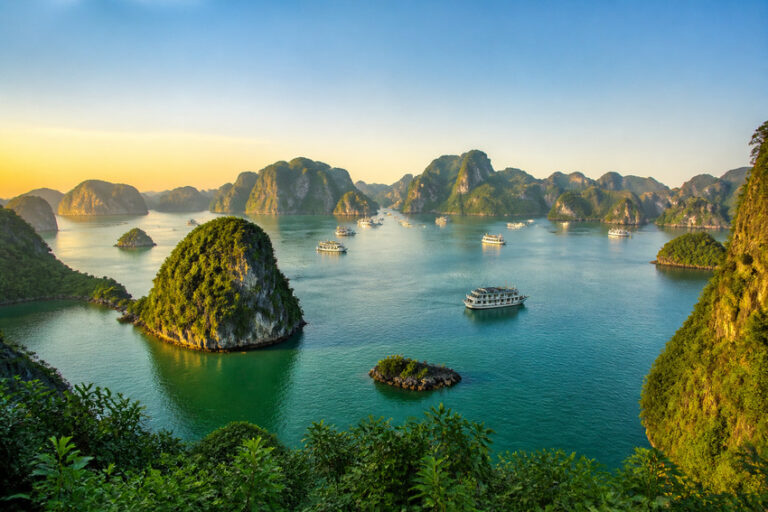 Aerial view of Ha Long Bay-style limestone islands in turquoise water with boats and a sunset glow on the horizon
