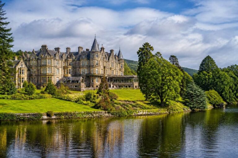 A large stone castle-style mansion on a grassy lakeshore with trees, reflected in calm water under a partly cloudy sky
