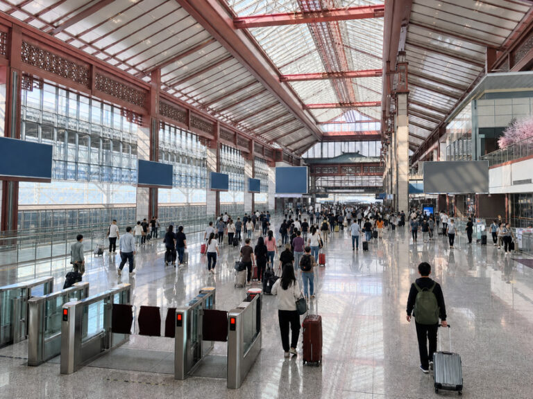 Busy airport terminal concourse with travelers and rolling luggage beneath a glass roof and red steel beams.