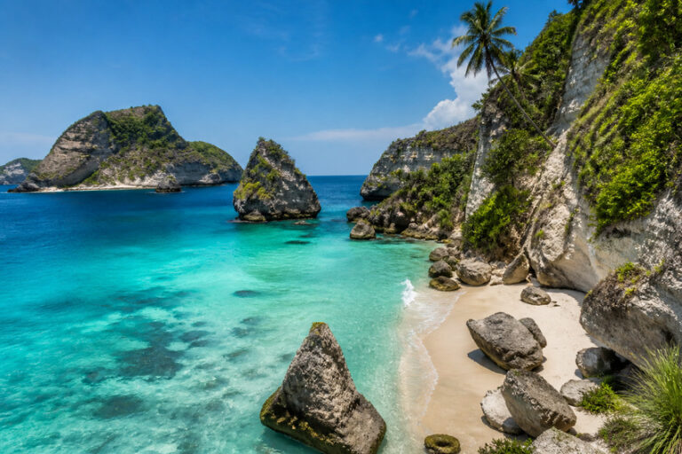 Turquoise water hugs a sandy beach surrounded by large rock formations and cliffs on a tropical island.