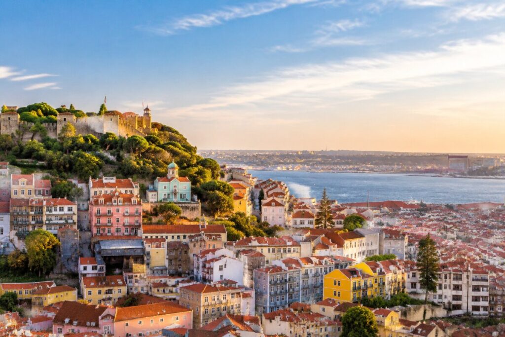Colorful hillside city by the water with a historic stone wall and castle on a hill at sunset‑lit coast.