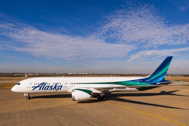Alaska Airlines passenger jet parked on a taxiway with a blue sky overhead and a teal-green tail design