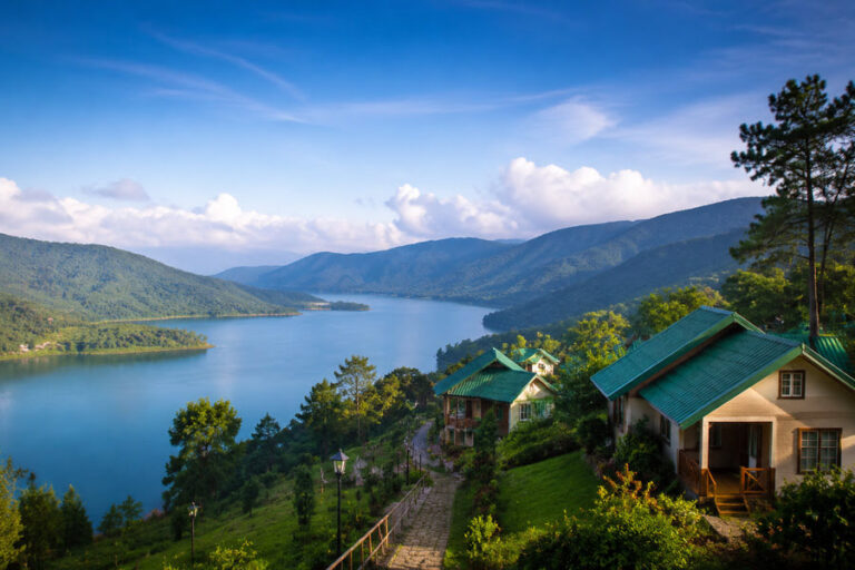A tranquil blue lake winds through forested mountains, with green-roof cottages on a hillside in the foreground.
