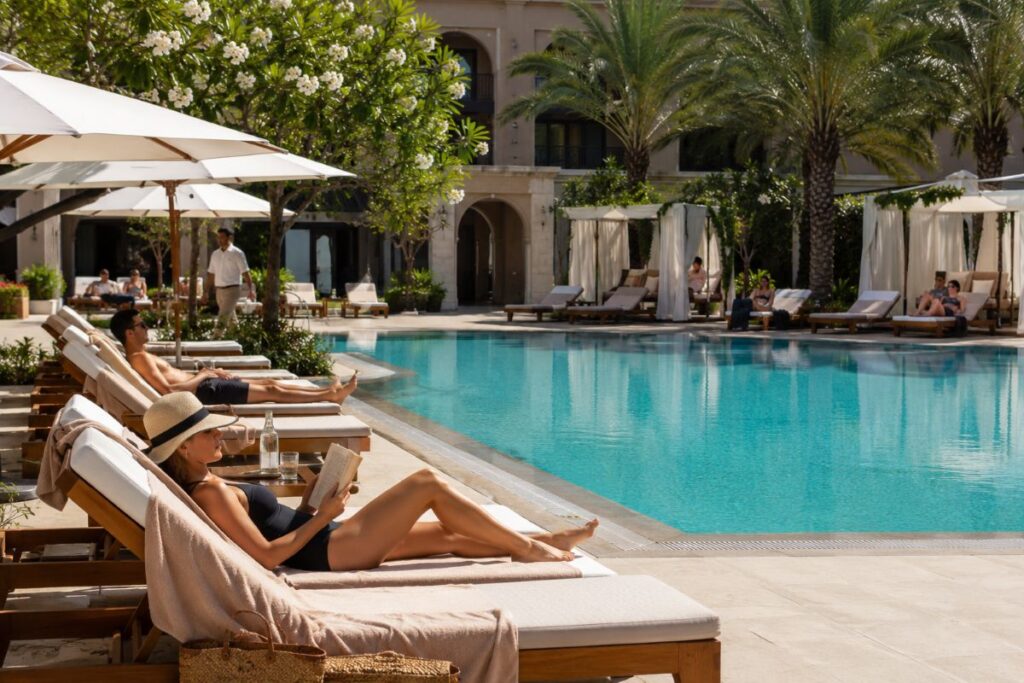 Poolside at a resort with lounge chairs, umbrellas, and palm trees on a sunny day.