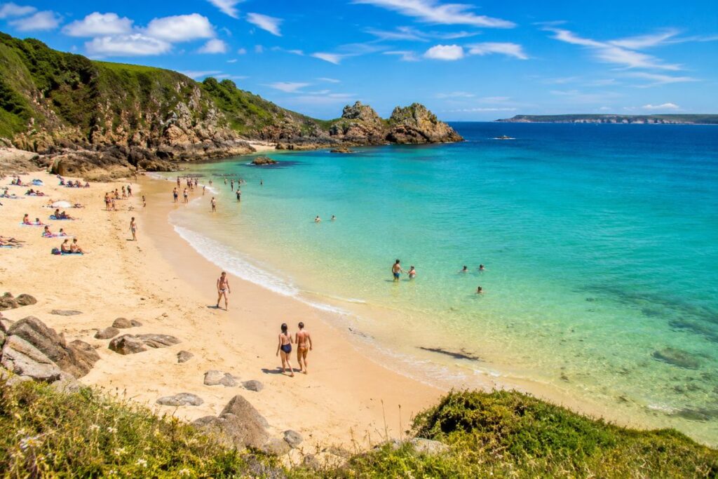 Sunny sandy beach with turquoise water, rocky cliffs, and people sunbathing and swimming on a bright day.