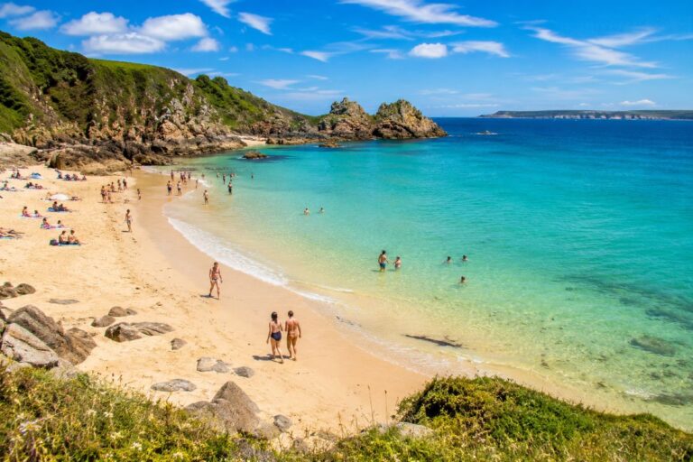 Sunny sandy beach with turquoise water, rocky cliffs, and people sunbathing and swimming on a bright day.