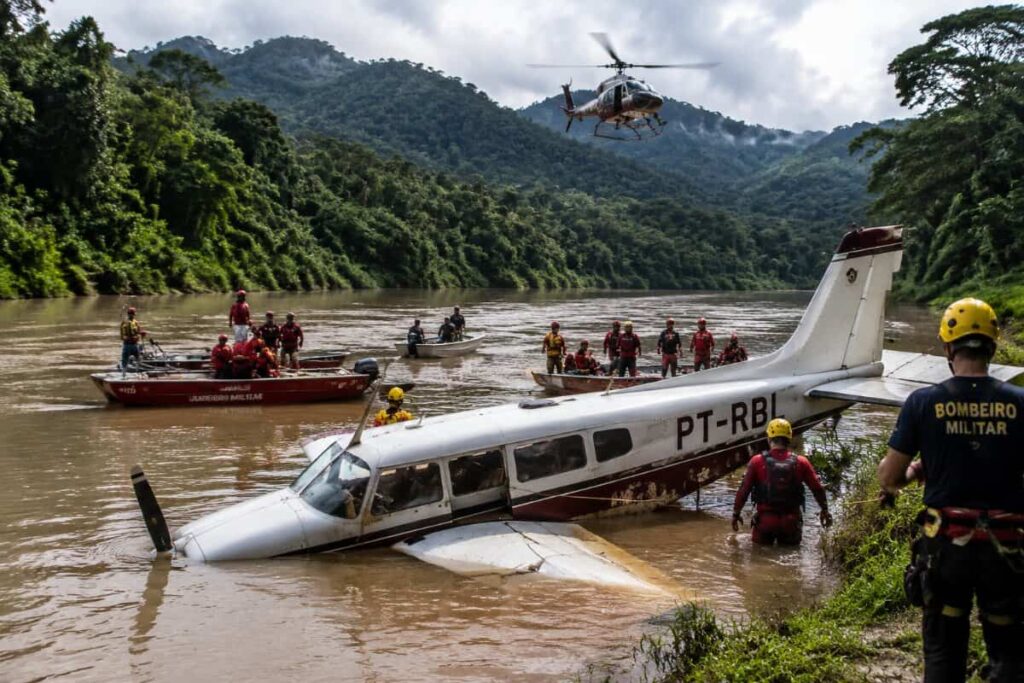 Rescue scene by a muddy river with a sunken small airplane titled PT-RBL, rescuers in red gear and boats nearby, helicopter overhead.