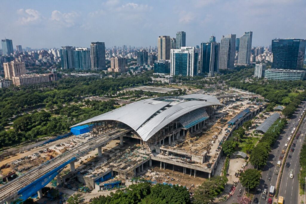 A large elevated railway station under construction with a curved white roof in a dense urban area.