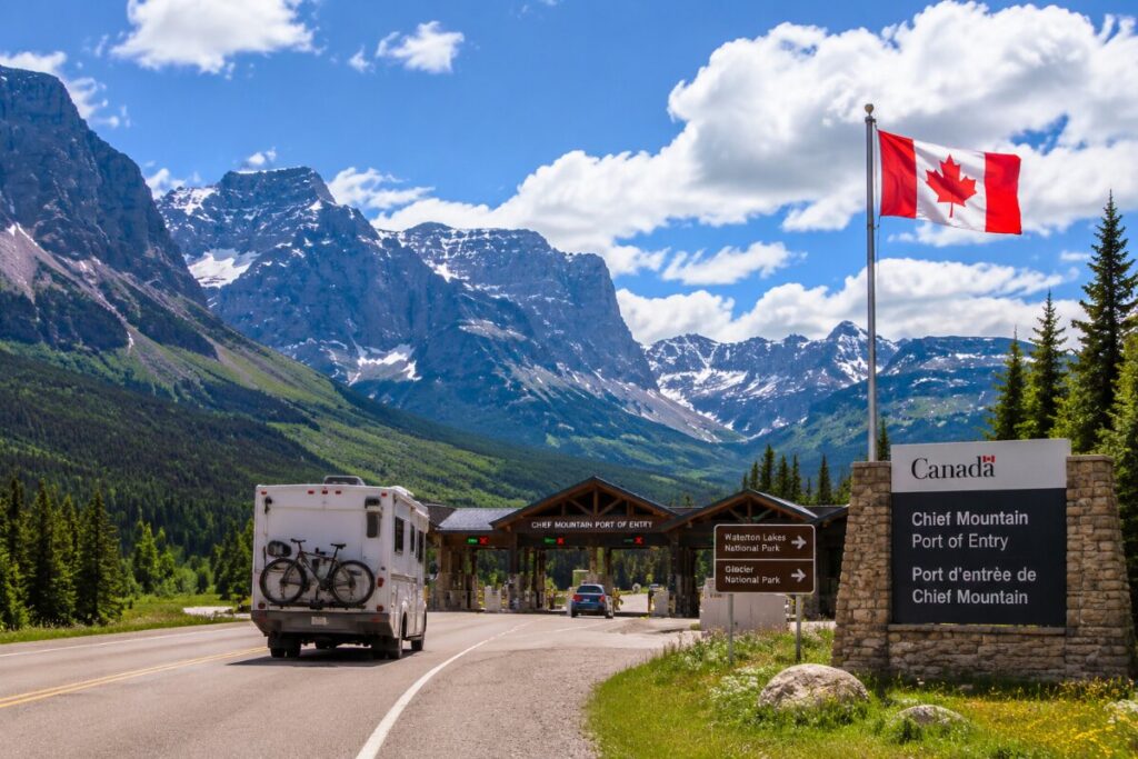 Canadian flag flying at Chief Mountain Port of Entry as a camper van with a bike on back approaches the border, set against snow‑capped mountains and pine trees.