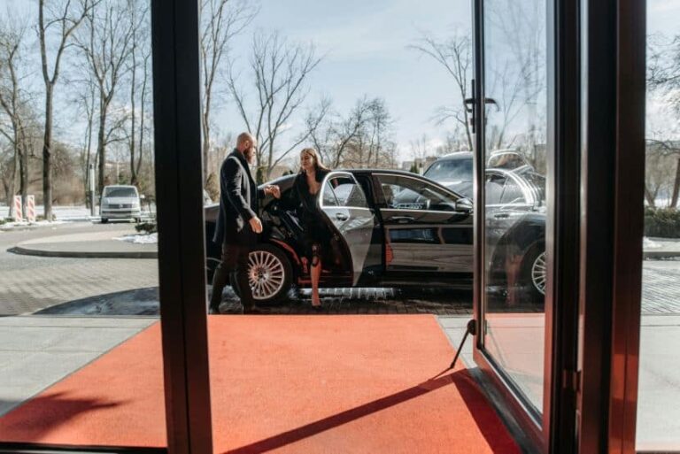 Man in a suit helps a woman exit a black luxury car onto a red carpet outside a glass-door entrance.