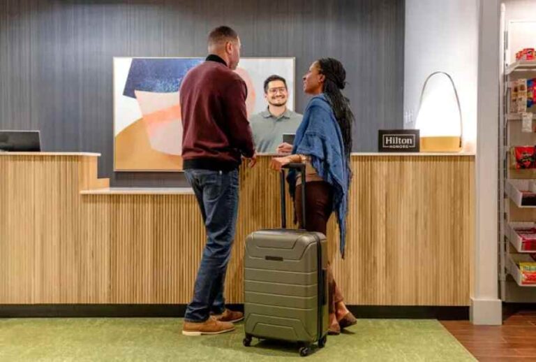 Man and woman with a rolling suitcase talk to a hotel front desk clerk at a reception area. Featuring a Hilton sign and a staff member smiling in the background.