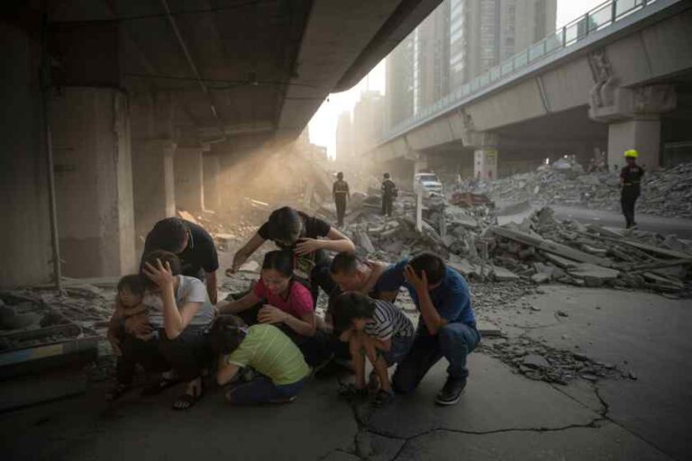 A group of adults and children huddles among rubble under a concrete overpass after a collapse, dust filling the air with distant workers in the background.