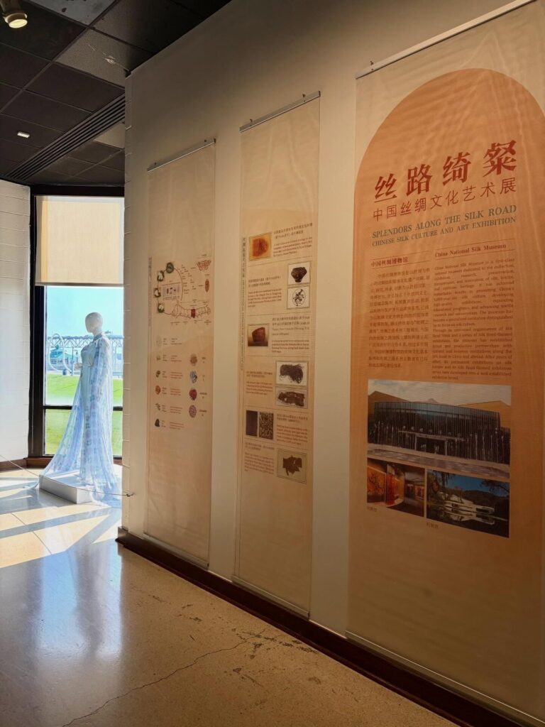 Exhibit wall with tall vertical panels of information; a pale blue translucent dress on a mannequin near a large window.