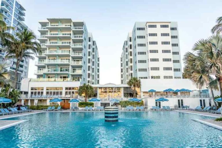 Resort hotel with a large outdoor pool, palm trees, and two mid-rise buildings in the background from a sunny vantage point.