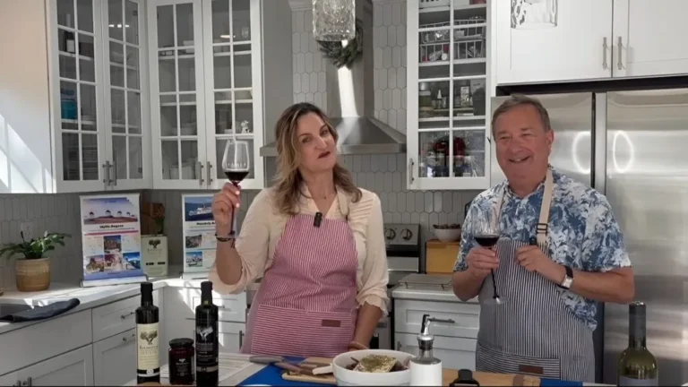 Two hosts in a modern kitchen toast with red wine during a cooking show or demo.