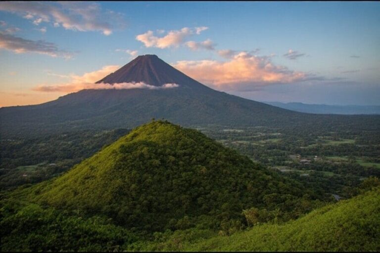 Cone-shaped volcano rising above forested hills under a pastel sunset sky.