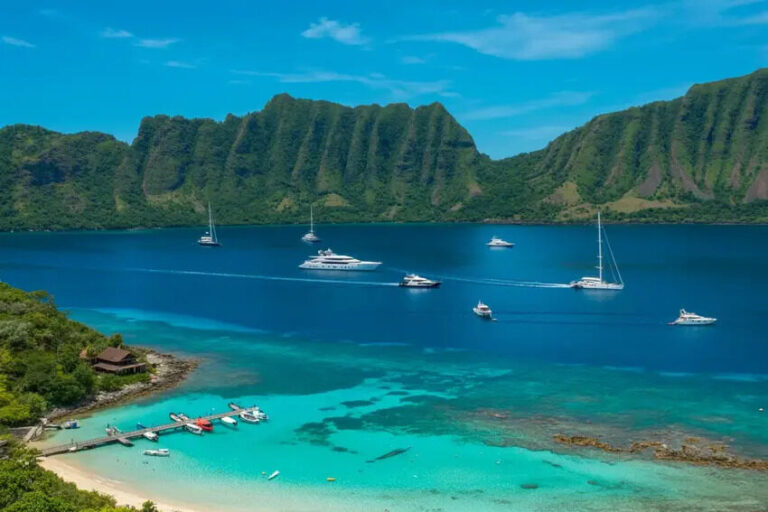 Turquoise bay with a white-sand beach, lush green cliffs, and several anchored boats.