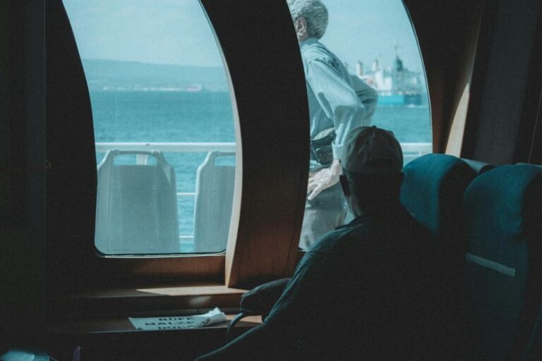 Man in a cap seated inside a ferry cabin, looking toward the water through curved windows.