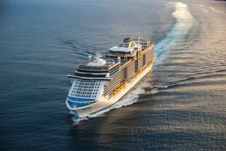 Large cruise ship sailing through calm blue water, leaving a white wake behind it, with multiple decks and balconies visible.