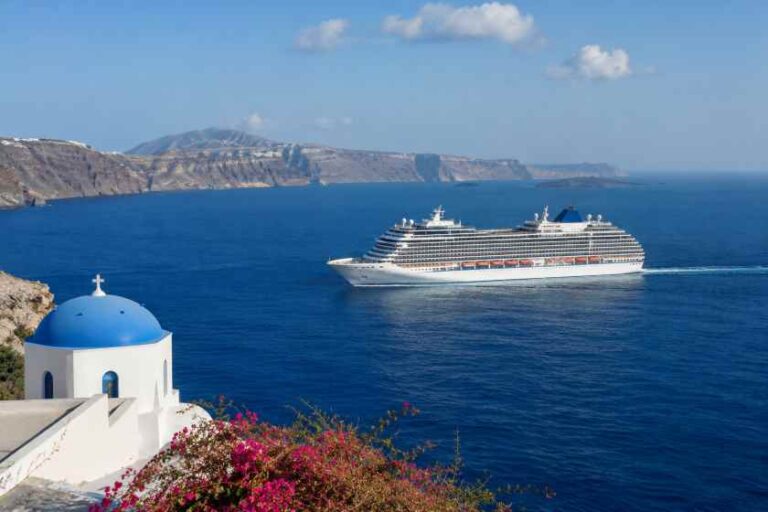 Blue-domed chapel on a cliff overlooking the Aegean Sea as a large cruise ship sails by.