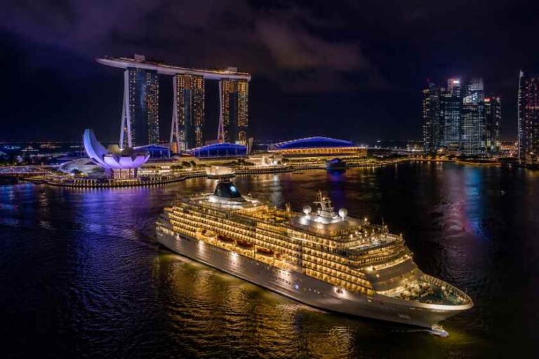 Nighttime view of a large illuminated cruise ship docked in Singapore's Marina Bay, with the Marina Bay Sands towers and city lights in the background.