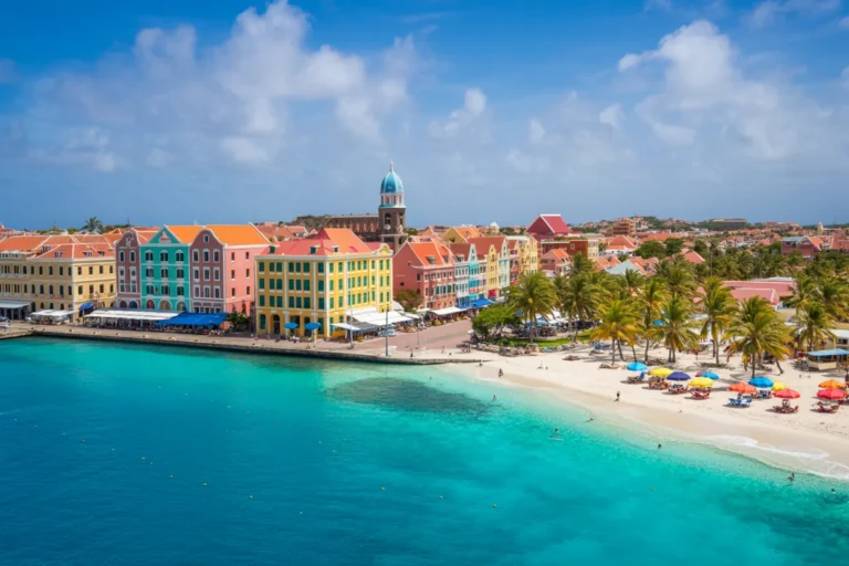 Colorful waterfront of a Caribbean town with pastel buildings, turquoise water, and a sandy beach with umbrellas.