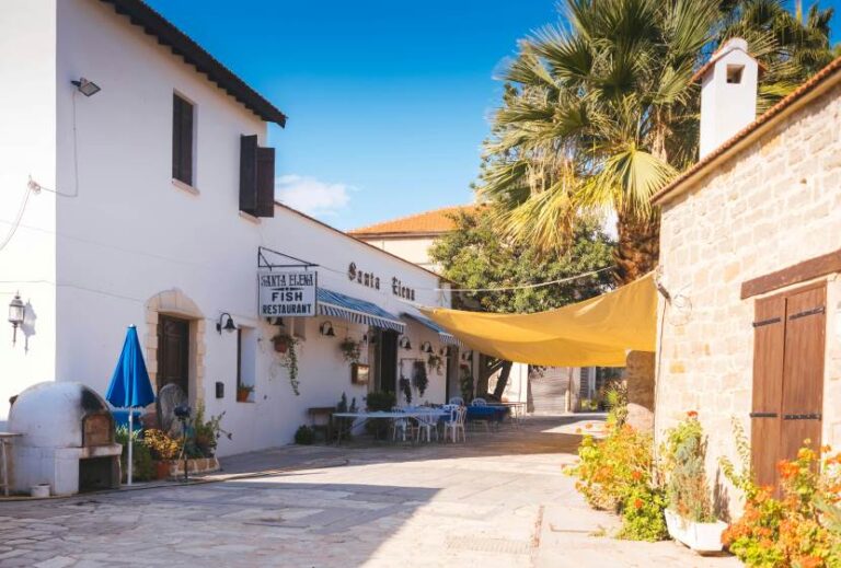Outdoor seating at Santa Elena Fish Restaurant along a sunlit white building, with a yellow canopy and palm trees nearby.