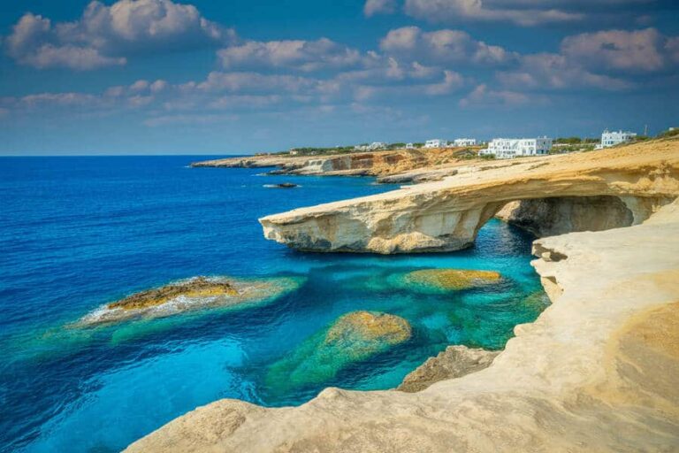 Turquoise sea beneath a rocky coastal arch with white buildings on the horizon on a sunny day.