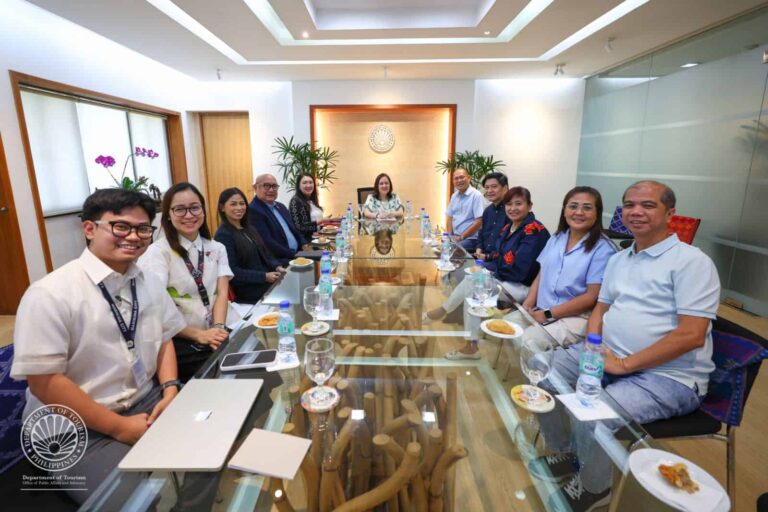 Group of professionals seated around a long glass conference table in a bright meeting room; Department of Tourism seal visible in the lower left corner.