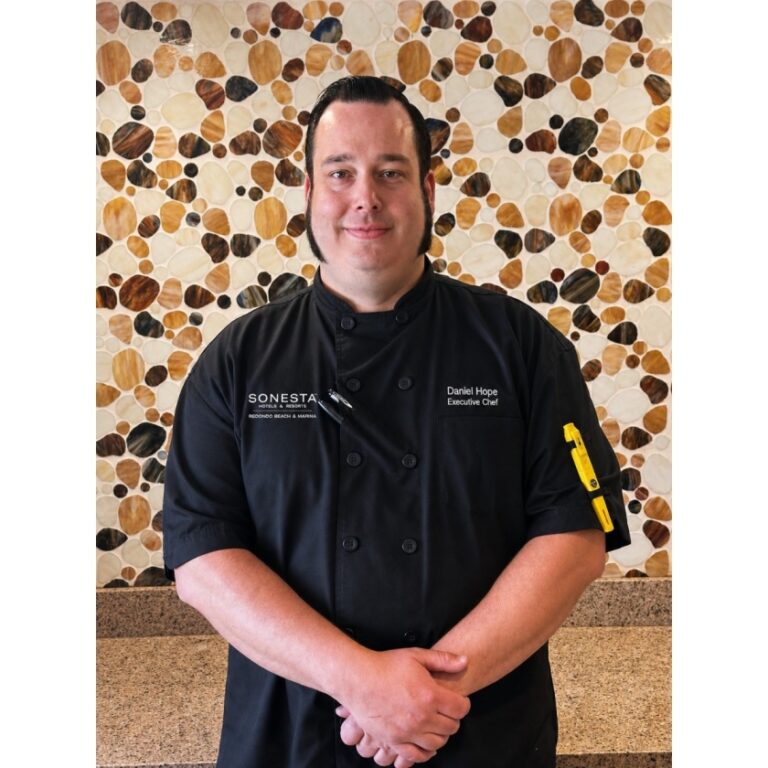 Executive chef Daniel Hope in a black chef coat, hands folded, standing in front of a pebble mosaic wall.