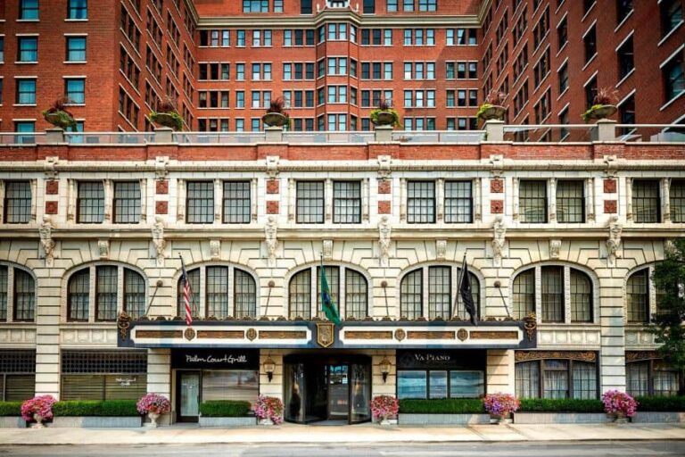 Historic white stone building with arched windows and a decorative storefront entrance beneath flags and planters along the sidewalk Chicago-style facades behind them.