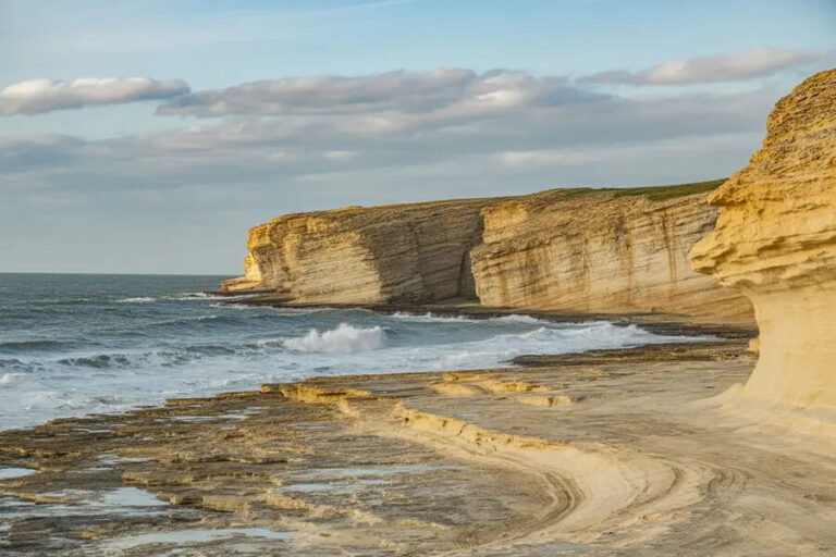 Sunlit limestone cliffs along a rocky shoreline with waves crashing at the base.