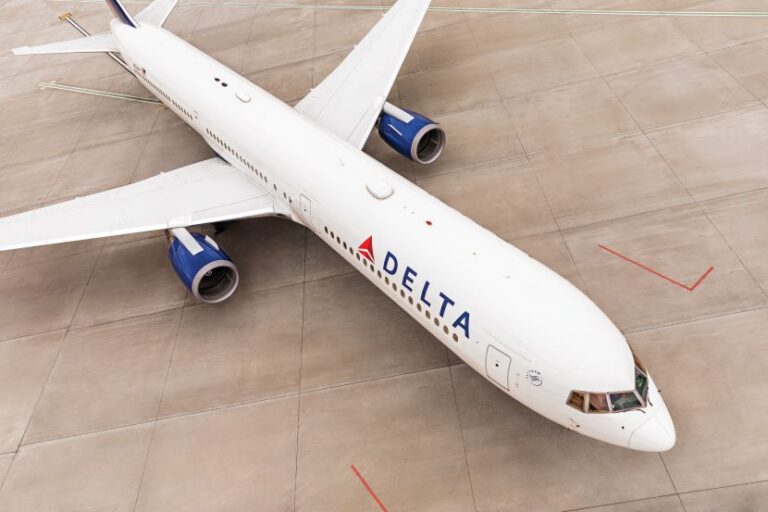 Top-down view of a white Delta Air Lines aircraft parked on the tarmac, blue engine nacelles under the wings, Delta livery visible on the fuselage.