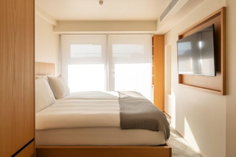 Hotel room with a large bed, white linens, gray throw, and a wall-mounted TV on wood paneling opposite the bed.