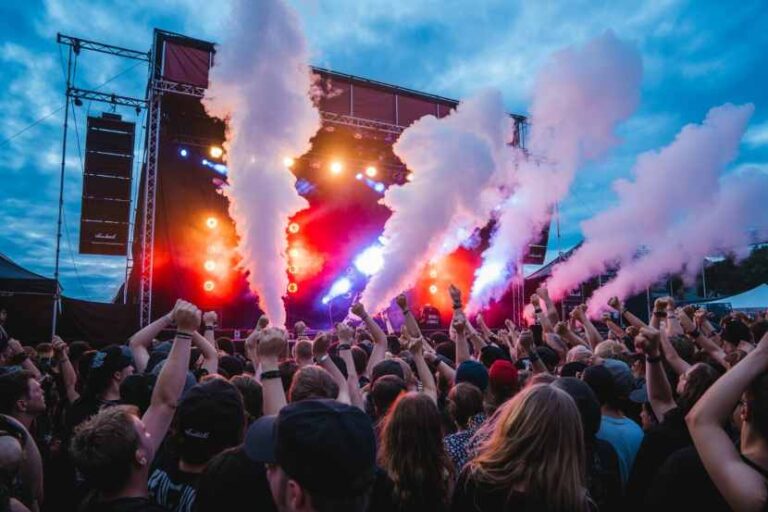 Crowd at an outdoor concert with a lit stage and colorful smoke plumes.