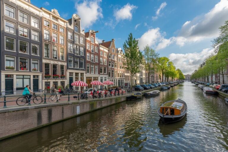 Colorful canal-side street in Amsterdam with tall gabled houses, cyclists, and a cafe with red umbrellas.