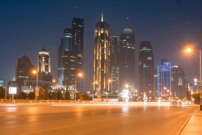 Nighttime city skyline with tall, illuminated skyscrapers and a brightly lit street in the foreground.