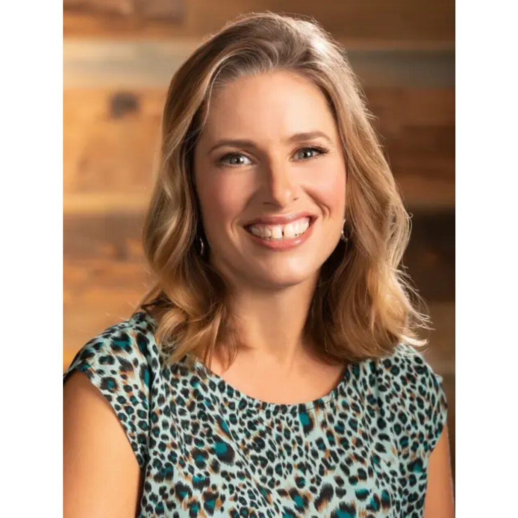 Smiling woman with shoulder-length blonde hair wearing a teal leopard-print blouse, in a warm wooden indoor setting.
