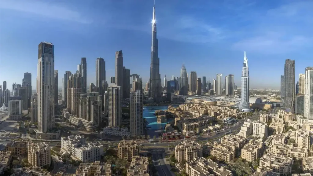 Panoramic city skyline featuring numerous high-rise towers under a clear blue sky, with a tall central skyscraper as the focal point (likely Burj Khalifa).