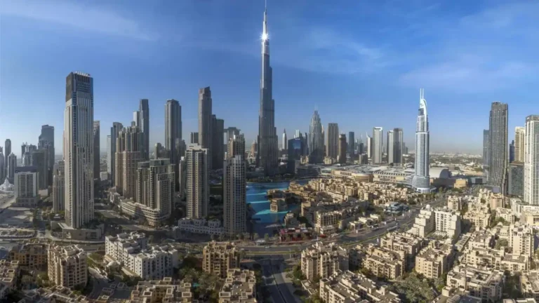 Panoramic city skyline featuring numerous high-rise towers under a clear blue sky, with a tall central skyscraper as the focal point (likely Burj Khalifa).