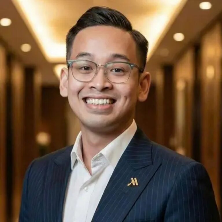 Smiling man in a navy pinstripe suit and white shirt, wearing glasses, in a warmly lit hallway or lobby.