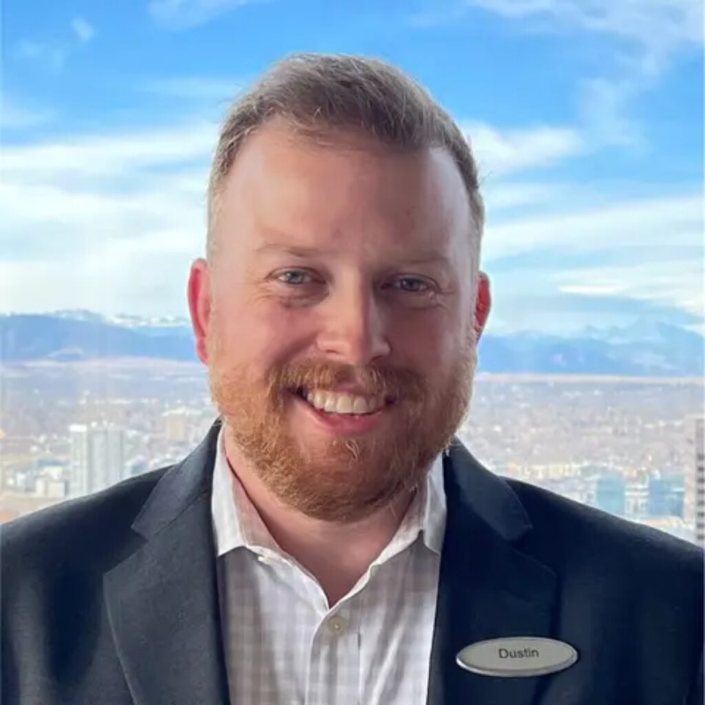 Smiling man in a dark blazer and white shirt, wearing a name badge that reads Dustin, with a city skyline behind him.
