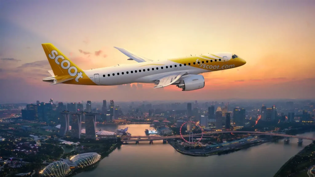 Scoot commercial airplane with a yellow tail flying over a city skyline at sunset.