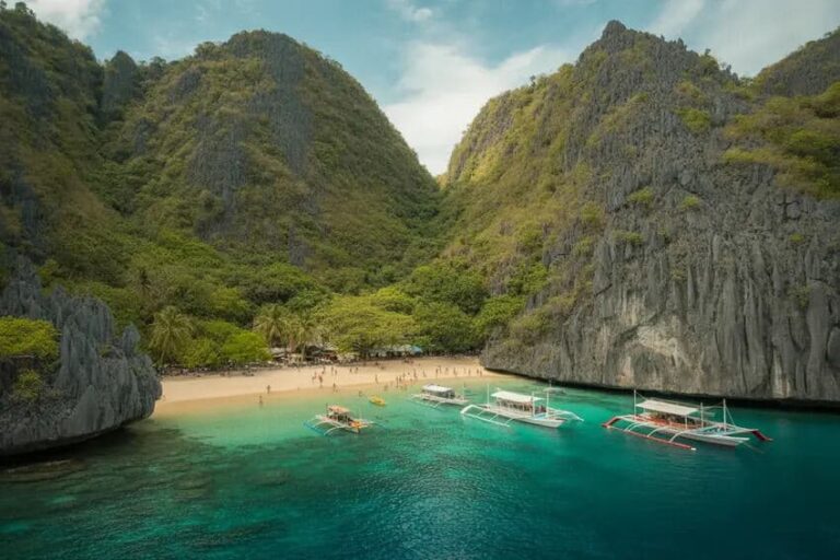Turquoise bay with white-sand beach nestled between towering limestone cliffs; colorful outrigger boats anchored near shore and a few beachgoers visible